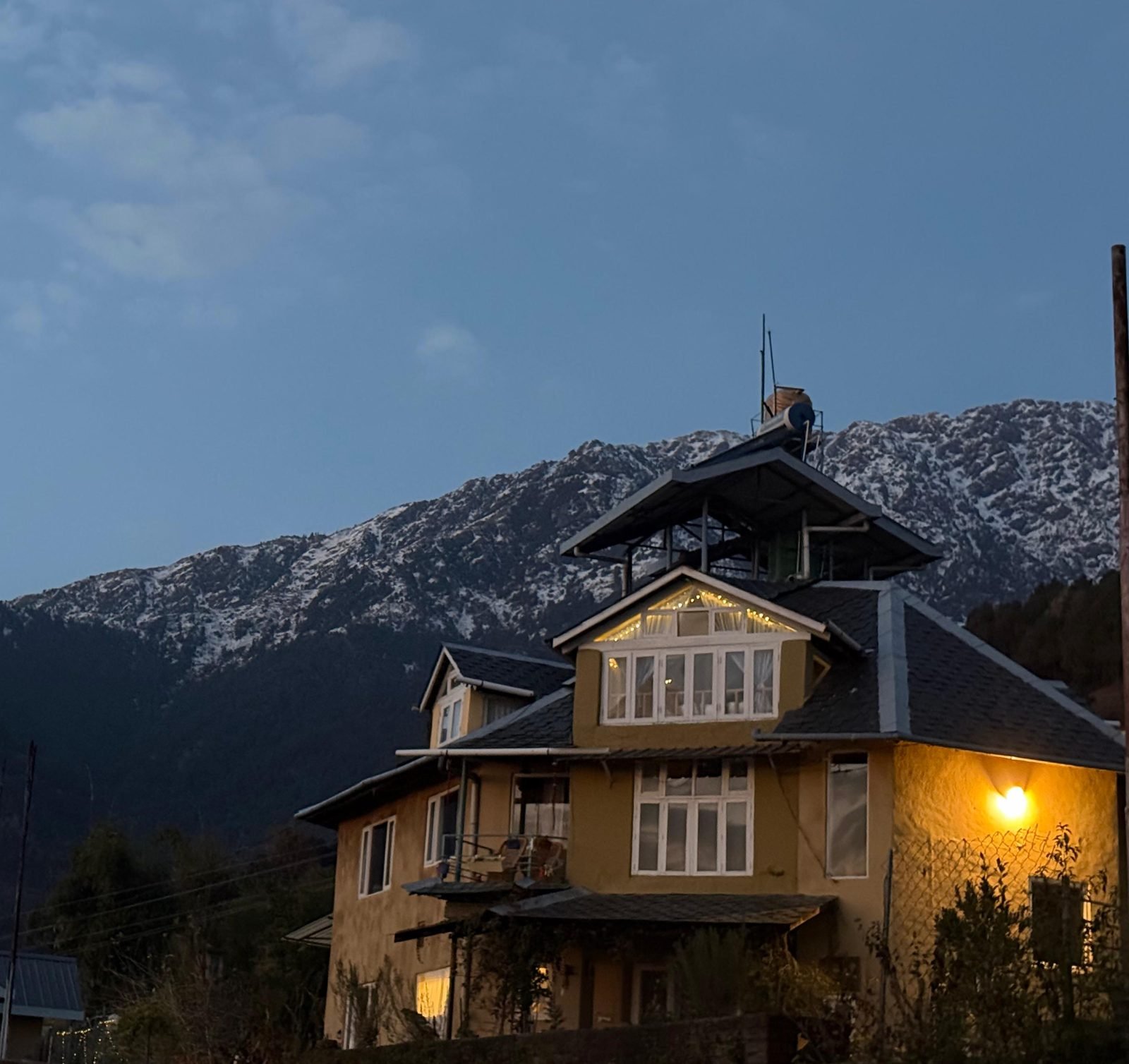 A hill house in Palampur at dusk, lights on, below the snow on the Dhauladhar